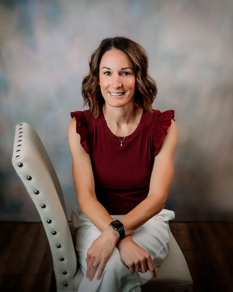 Melodie Look. White woman with shoulder length brown hair. Wearing a red blouse and white pants. She is sitting on a white chair.